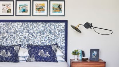 A white bedroom with double bed to the left with statement blue and white patterned headboard, and blue cushions patterned with koi fish. To the right of the headboard is a brown wood bedside table with a small plant, books, a small bowl, and a framed photograph. Above is a wall-mounted angle poise lamp, and above the bed are four small, square framed artworks.
