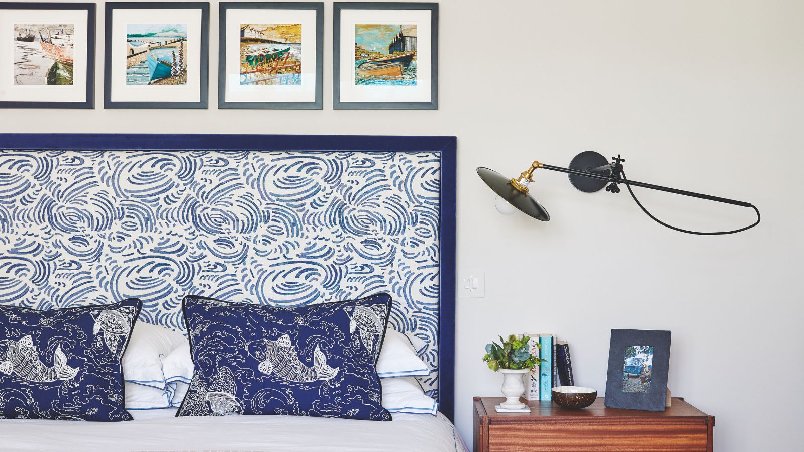 A white bedroom with double bed to the left with statement blue and white patterned headboard, and blue cushions patterned with koi fish. To the right of the headboard is a brown wood bedside table with a small plant, books, a small bowl, and a framed photograph. Above is a wall-mounted angle poise lamp, and above the bed are four small, square framed artworks.