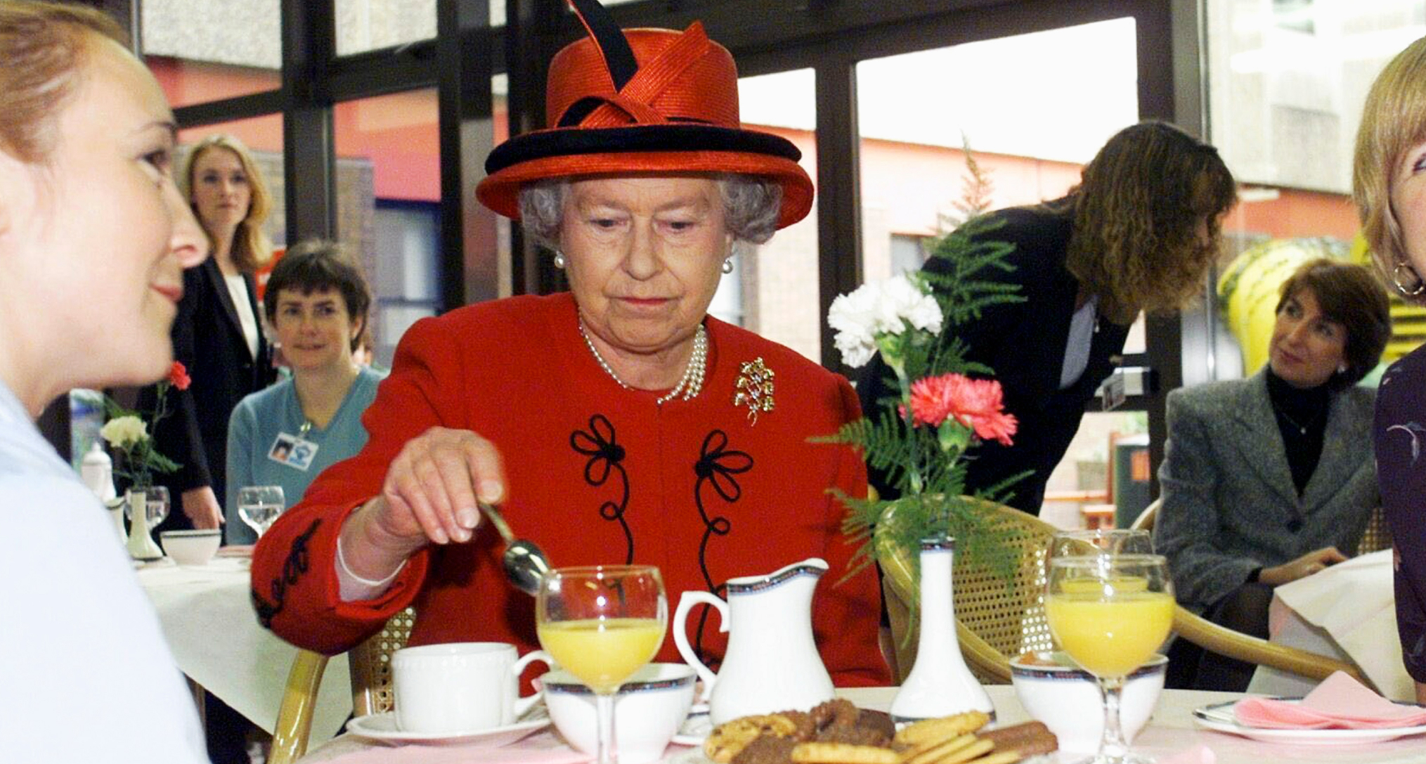 Queen Elizabeth in a red coat and hat stirring tea at a table