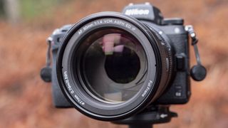 Close up of the Viltrox AF 85mm F1.4 Pro lens attached to a Nikon camera, mounted to a tripod outdoors and with bronze-colored ferns in the background, emphasis on the lens aperture blades
