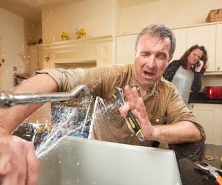 man being sprayed with water from leaking tap with wife in background on the phone