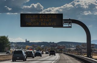 a line of cars stopped on a highway under a sign that reads "solar eclipse saturday. expect major delays"