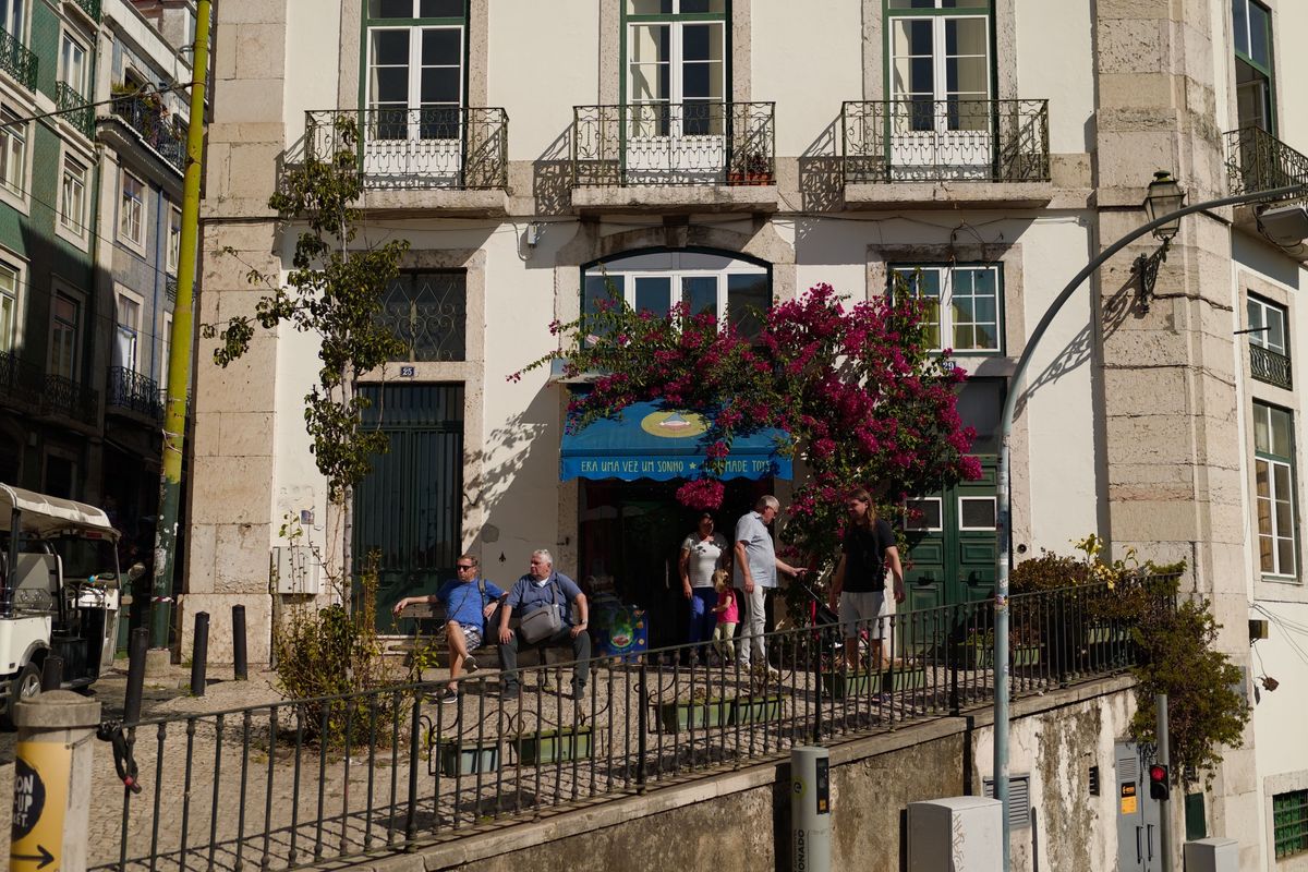 People gathered outside a white Lisbon building beneath balconies and bright pink bougainvillea in strong sunlight.