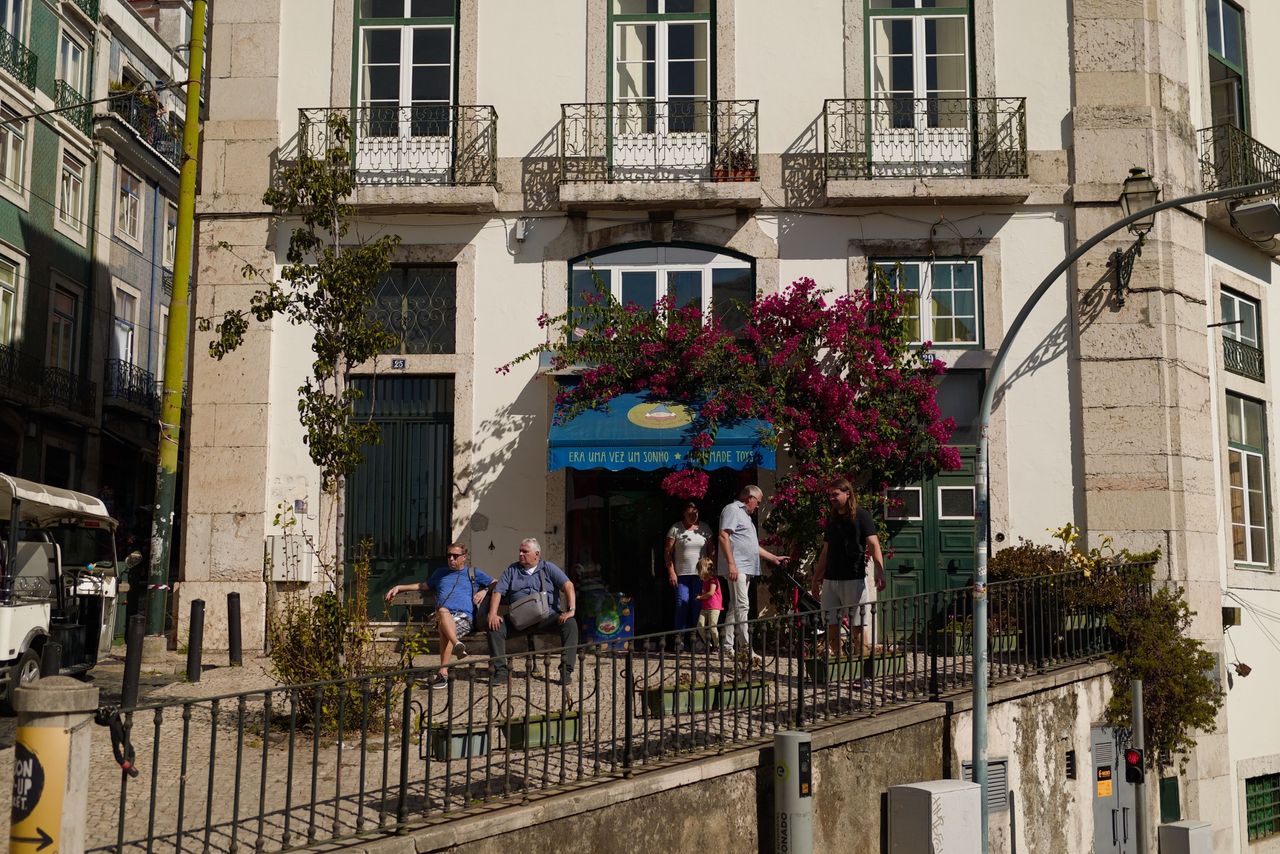 People gathered outside a white Lisbon building beneath balconies and bright pink bougainvillea in strong sunlight.