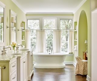 large bathroom with light yellow-green walls, white bath in front of window, vanity unit with double sinks and wooden floor