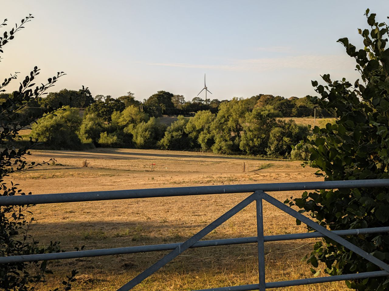 Metal farm gate between hedgerows overlooking dry fields and trees in the countryside at sunset.