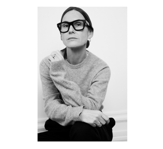 Louise Trotter wearing a crewneck sweater and glasses while standing in front of a blank wall