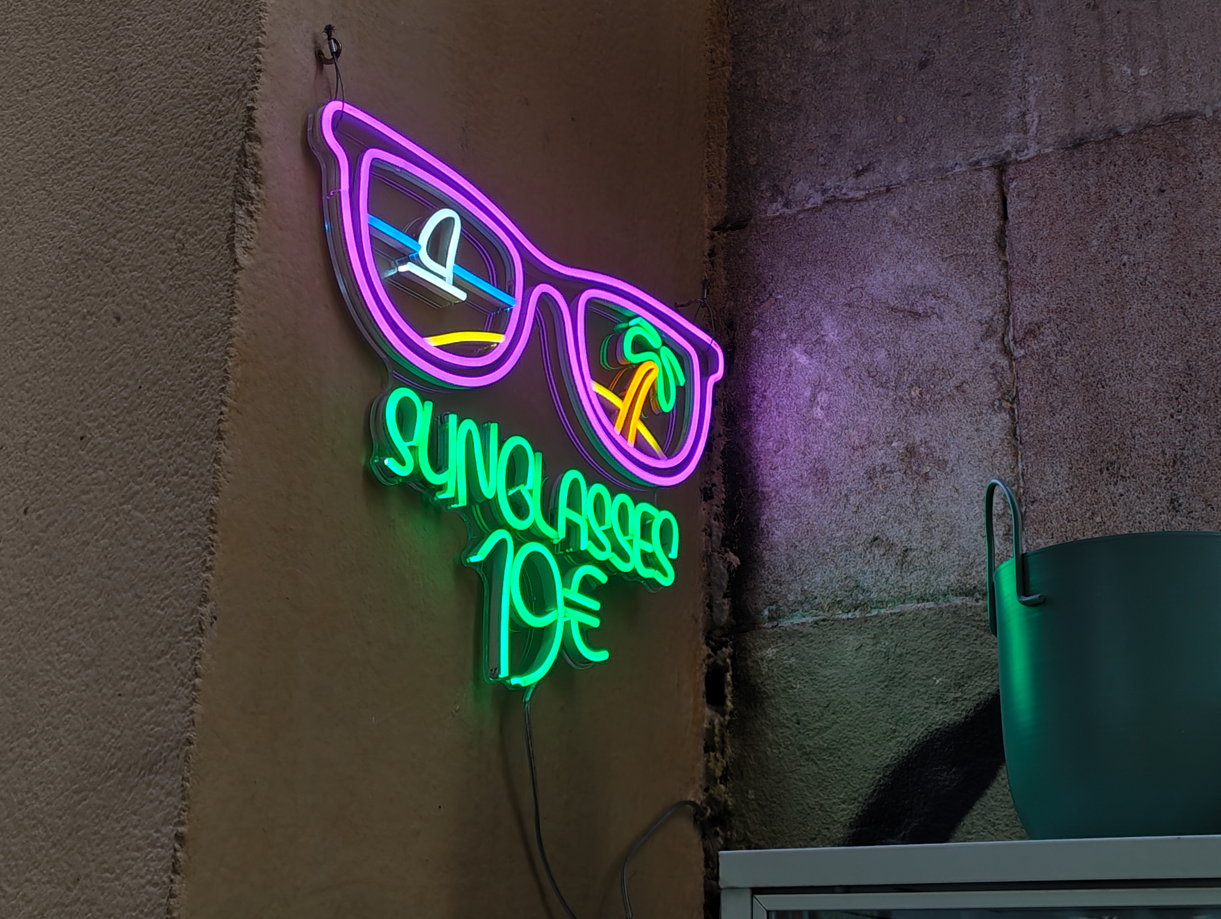 Neon sign shaped like sunglasses reading &ldquo;Sunglasses&rdquo; mounted on a textured wall, glowing in purple and green light, photographed with the Nothing Phone (4a).