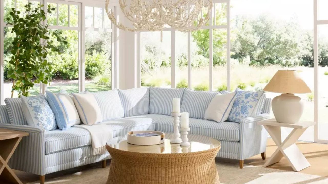 A blue sectional sofa covered with blue and white throw pillows behind a round rattan coffee table; a chandelier suspended from the ceiling; in the background, French doors looking onto a garden view.