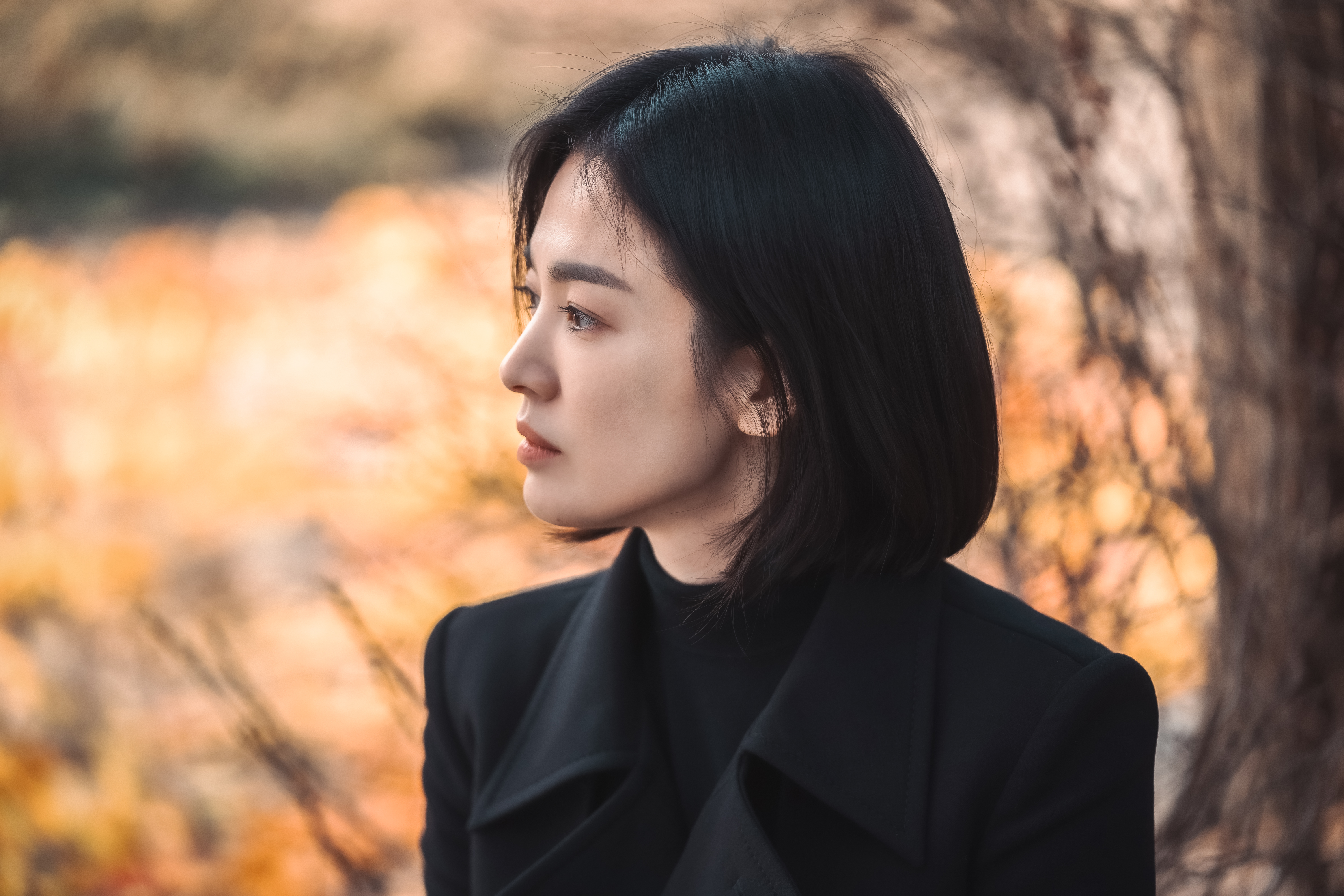 A Korean woman (Song Hye-kyo as Moon Dong-eun) wearing all black looks to the left as she stands outdoors, with a tree and fall leaves blurred behind her. A still from the K-drama 'The Glory.'