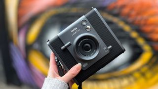 A hand holds up the Instax Wide Evo camera against a colourful graffiti wall