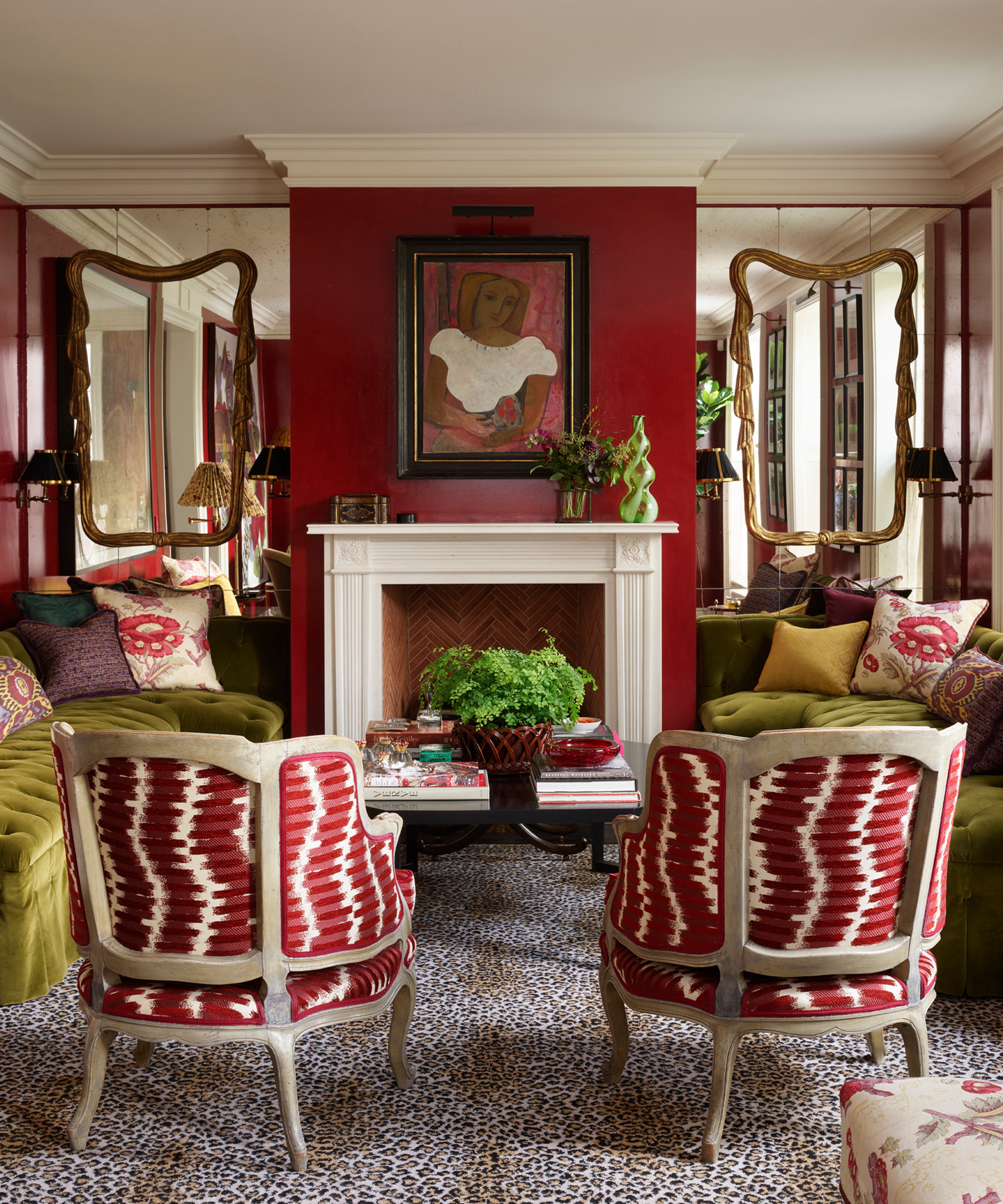 Bold living room featuring deep red walls, two olive green velvet sofas facing a fireplace, and two armchairs with red and white patterned fabric on a leopard print rug