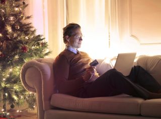 Mann i brun genser og svarte bukser bruker en laptop i en sofa.
