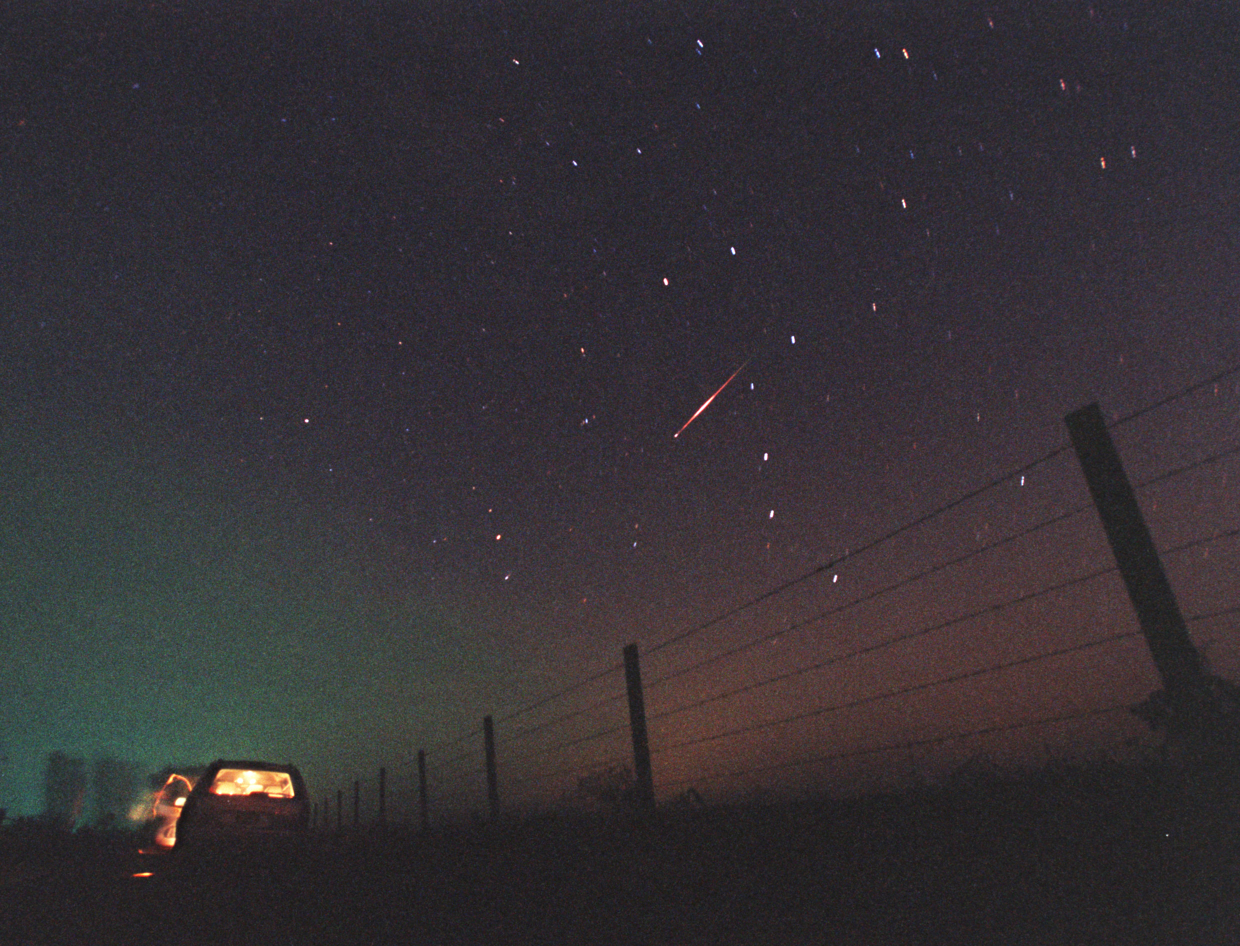 a streak of light in the starry night sky above a car parked next to a barbed wire fence