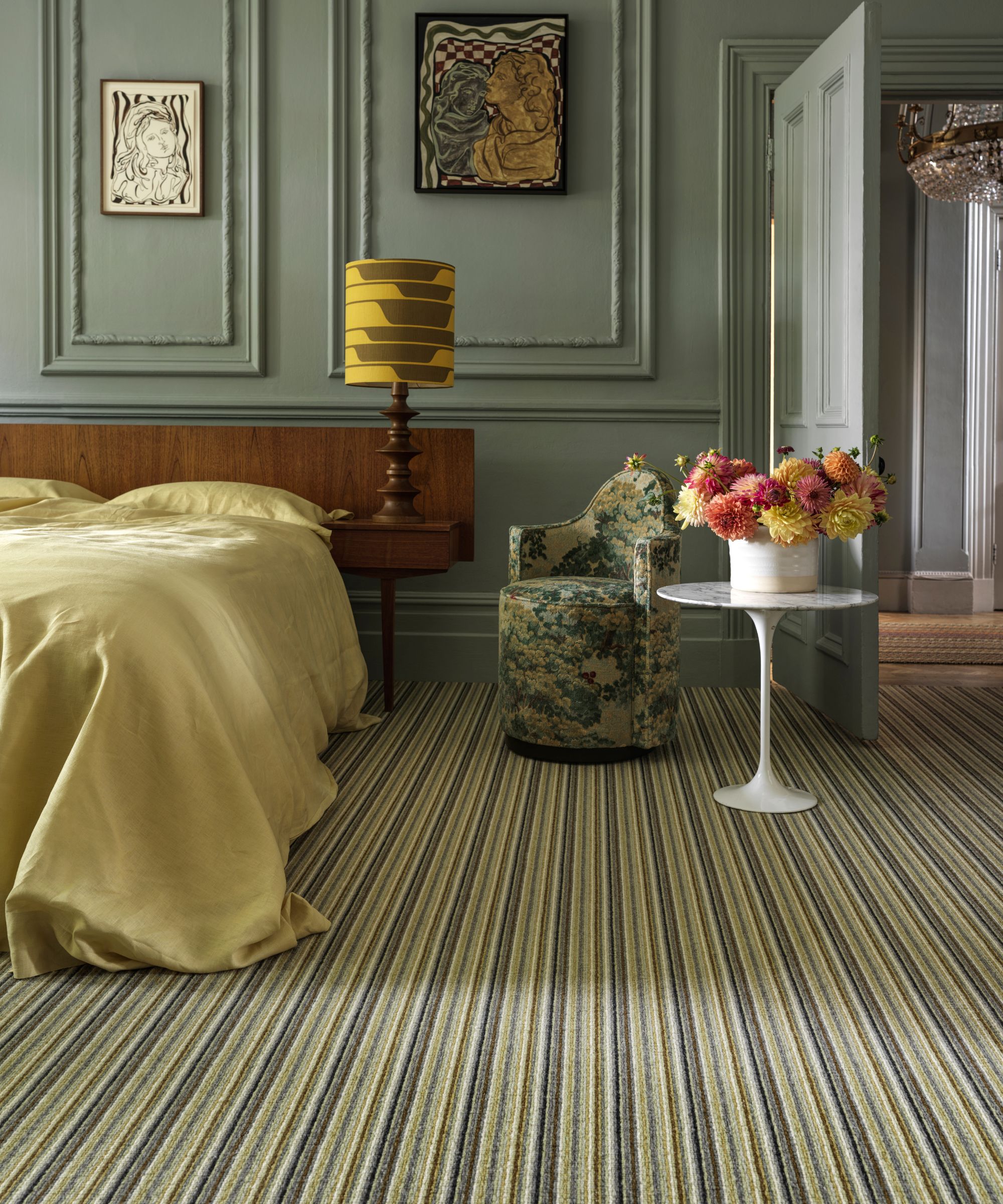 A modern bedroom with a green theme. Striped green carpet, a bed with gold-green bed linen, a modern wooden lamp with a mustard coloured lampshade, a small green chair and a white small table, on which is a white vase and many different coloured dahlias.