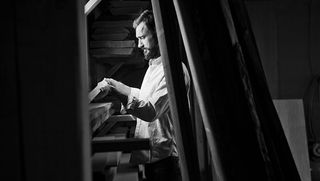 Black and white photo of Michael Moran holding a piece of wood at a shelving unit filled with wood planks