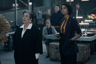 Wes (Caroline Quentin) and Archie (Anjli Mohindra) stand in the Lazarus Project office, which is a very grey and sombre environment. They are both looking up at an unseen screen high up on the wall with concern.