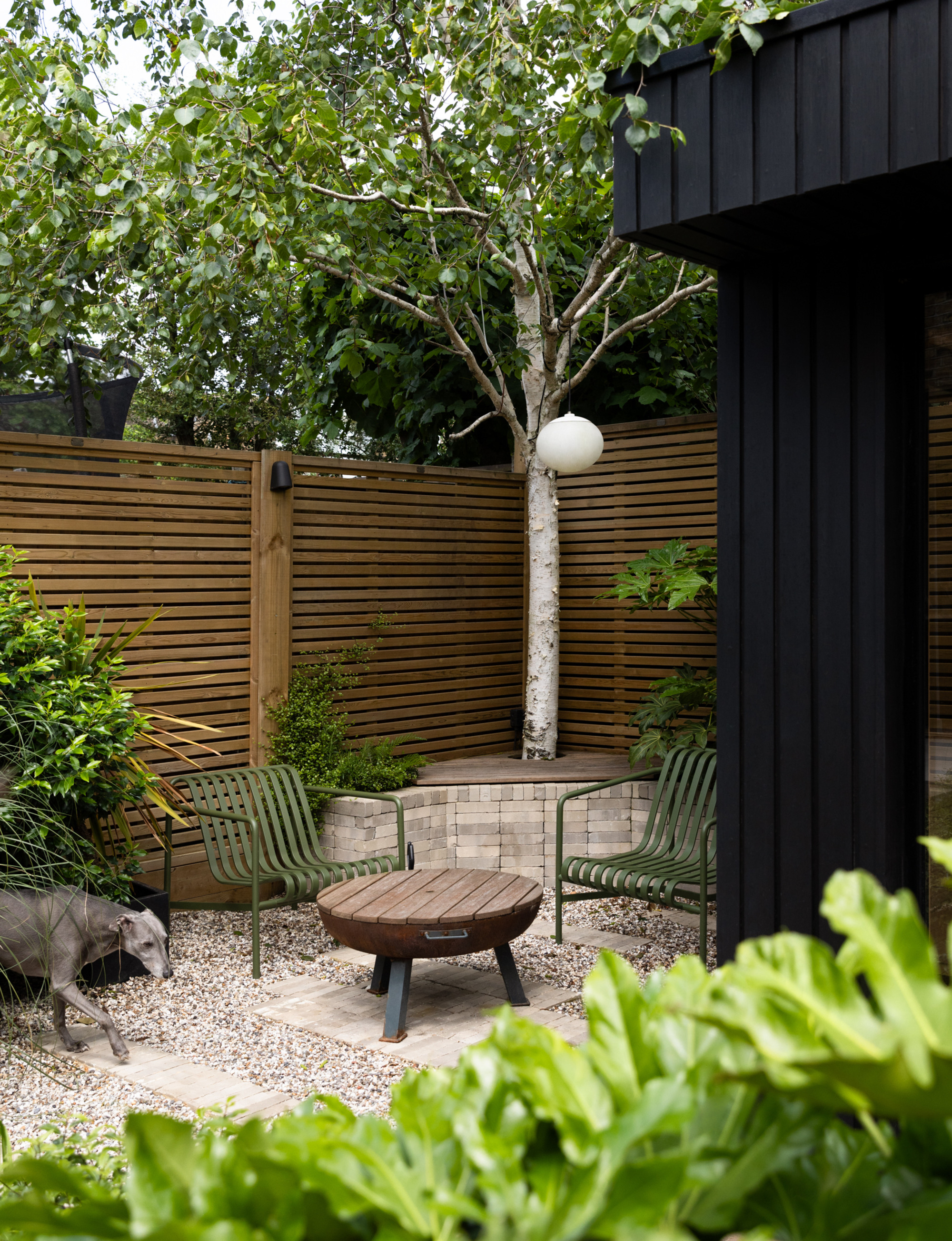 two green chairs beside a firepit in the corner of a garden with the firepit in the middle and a large tree placed at the back of the garden