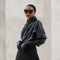 PARIS, FRANCE - DECEMBER 20: Emilie Joseph wears slicked-back hair held with a minimalist black headband, natural makeup with a smooth complexion and defined lips, black rectangular sunglasses, a vintage black leather jacket styled as a wrap top with a cinched waist creating a sculptural line, a black asymmetric pleated skirt by COS layered over black leggings, vintage black leather gloves, black leather slingback heels shoes with a square silver buckle by Saint Laurent, during a street style fashion photo session, on December 20, 2025 in Paris, France. (Photo by Edward Berthelot/Getty Images)