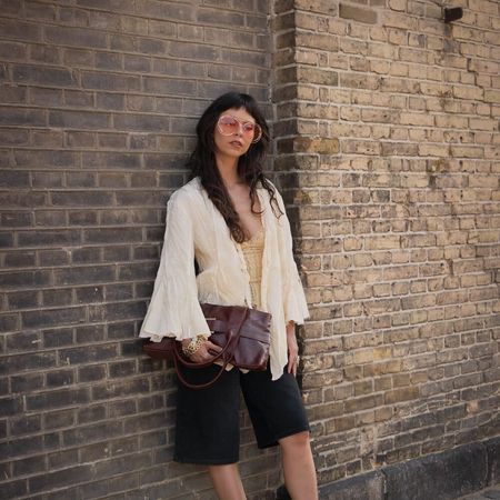 Woman wears white flowy blouse, black long shorts, tall square-toe boots, and a cute cluctch.
