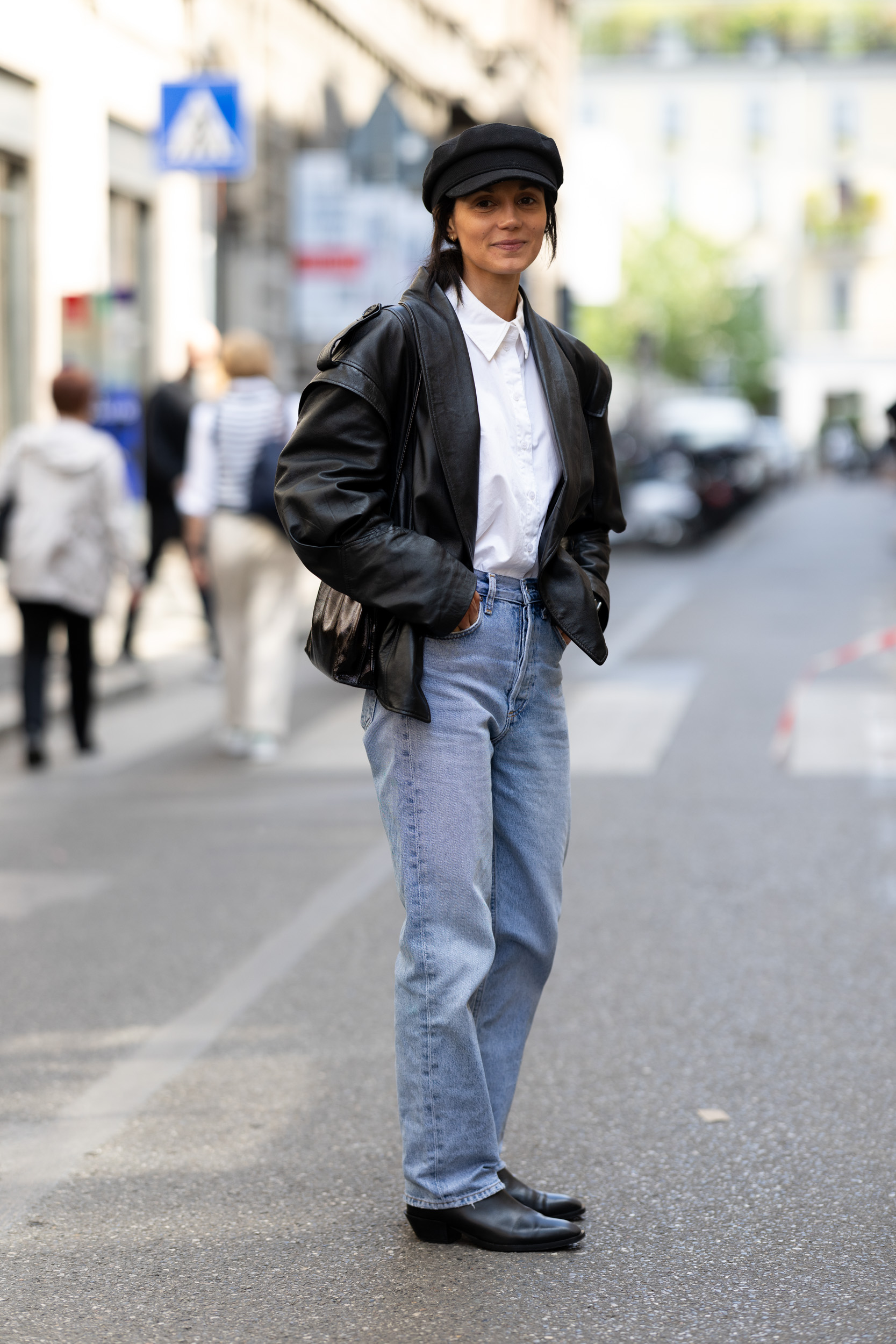 A woman wears a black hat, leather jacket, white tee, light-wash jeans, and black heels.