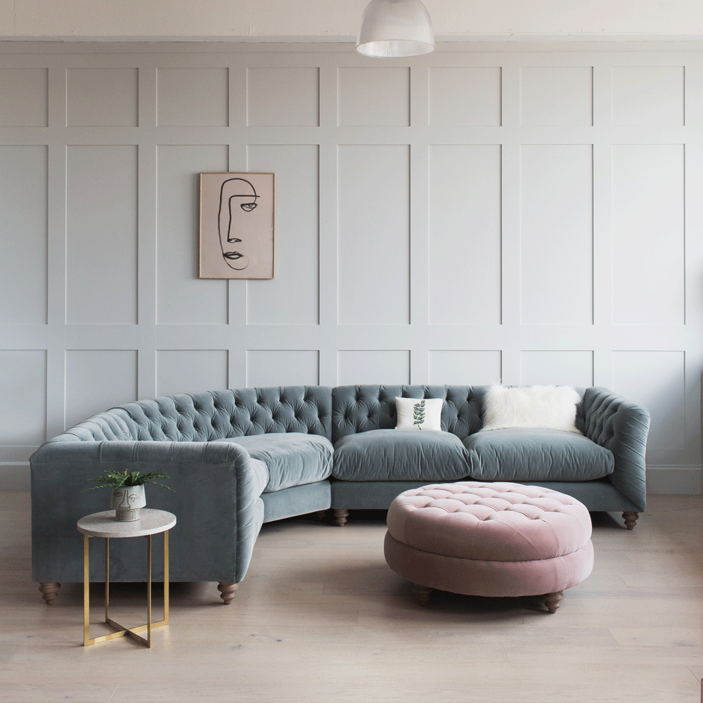 a blue-grey curved corner sofa and pink circular padded footstool, both with button detailing, a living room with light brown floor and light grey panelled wall