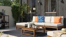 An outdoor seating area with a wooden garden sofa with cream cushions and a matching outdoor lounge chair and coffee table against a black-paneled wall with modern wall lights above