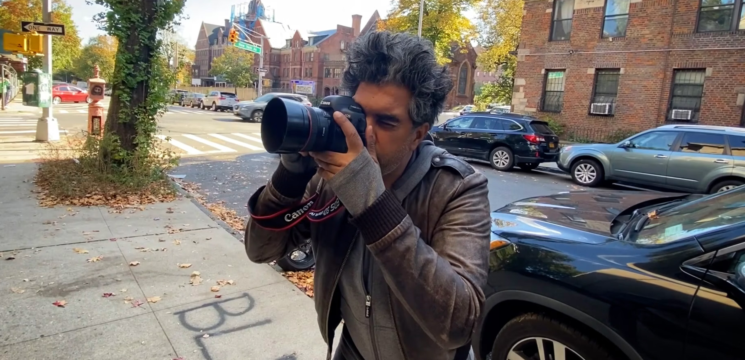 A man with dark curly hair raises a Canon DSLR camera to his eye on an autumn New York street, with brick apartment buildings