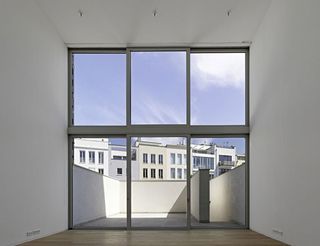 Interior room, wooden floor, white walls, window framed wall with view of white balcony and surrounding white buildings, blue sky with soft white clouds