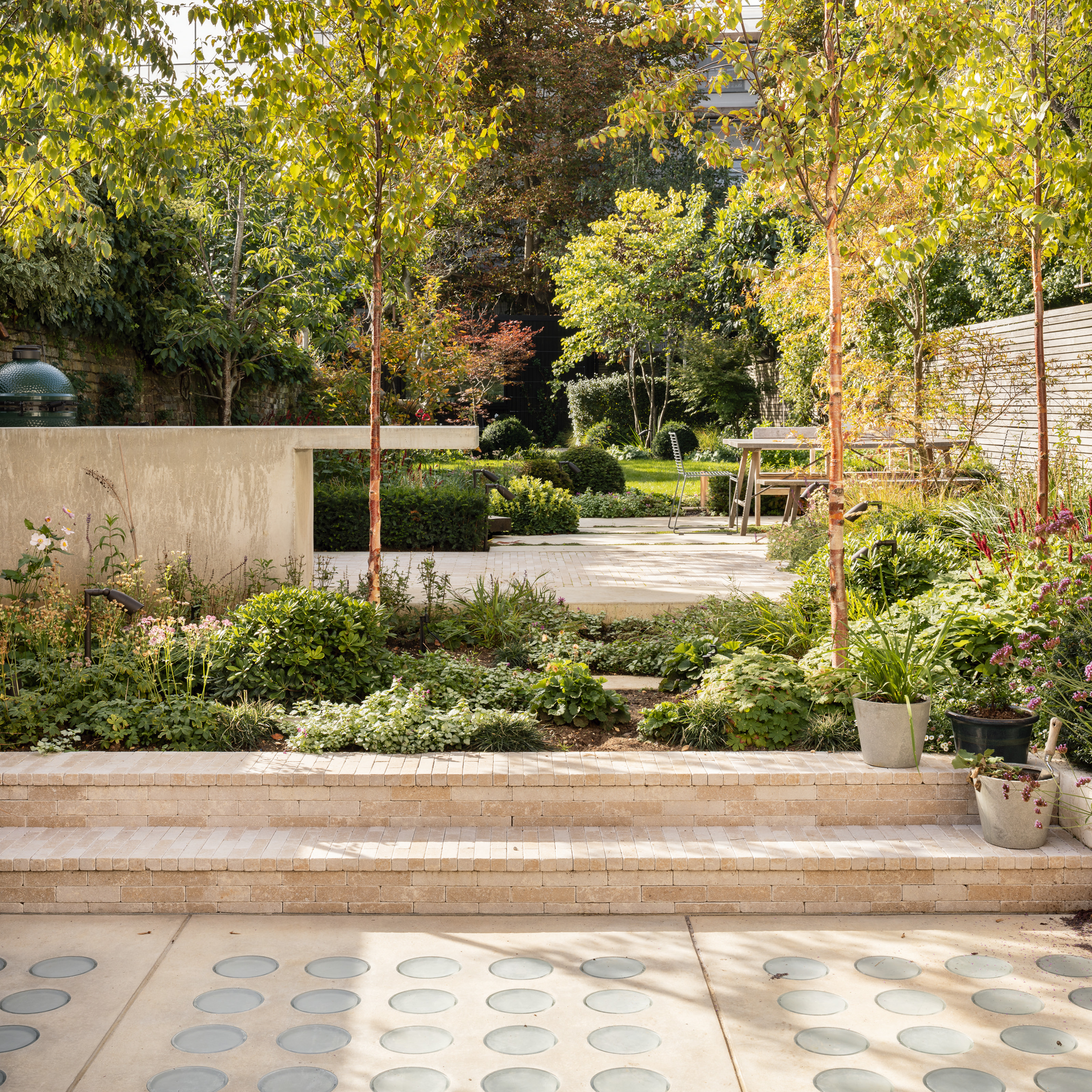 Tiered garden with pale stone and glass patio, pale brick steps and pale concrete outdoor kitchen