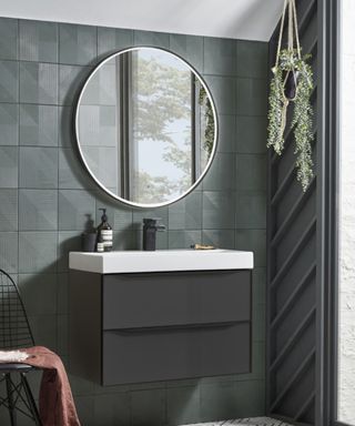 A bathroom with dark gray tiles, a circular silver mirror, a floating black sink with white surfaces, and a black chair to the left with a dusky pink blanket