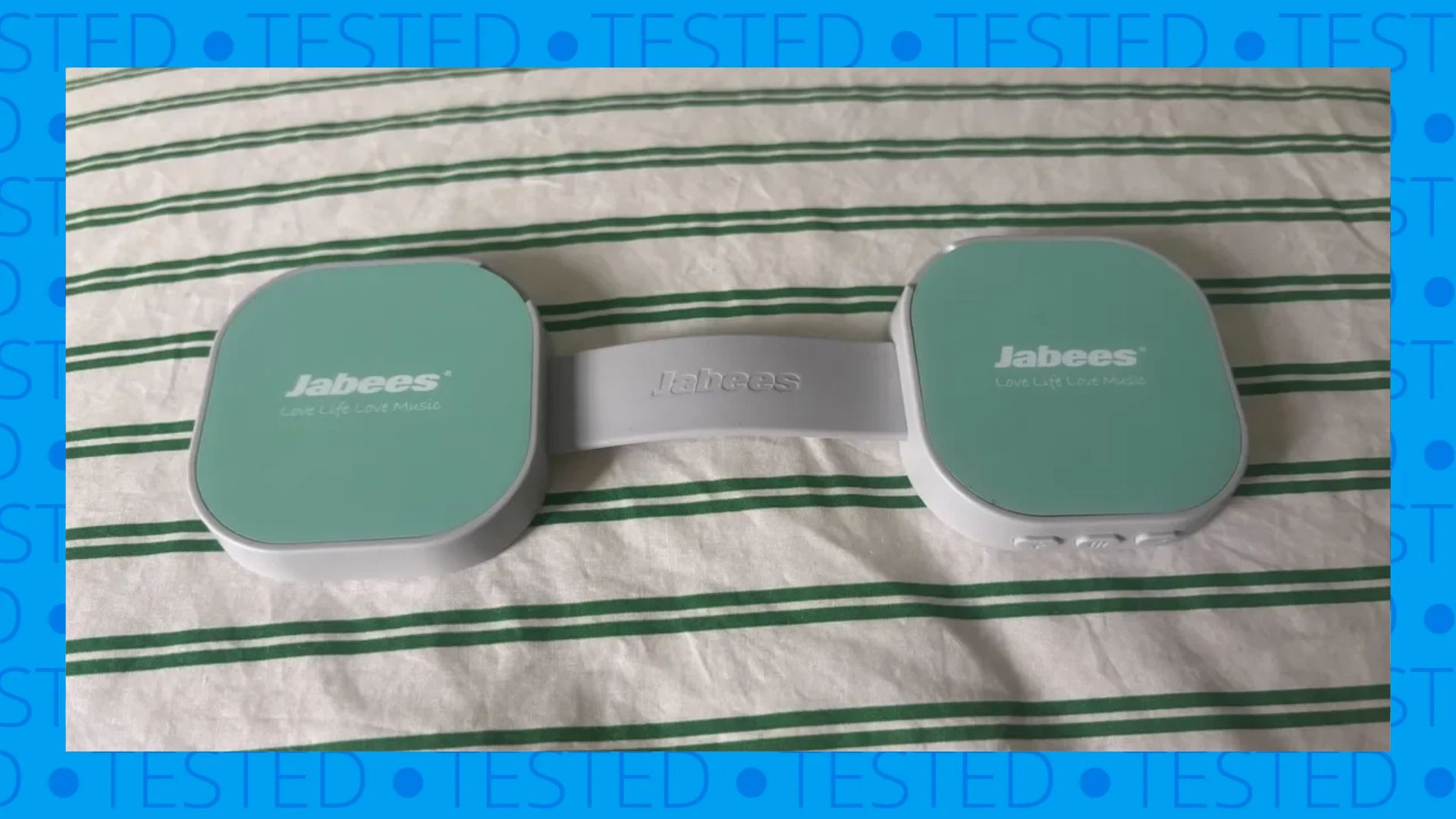 The image shows the flat green and white dumbbell-shaped Jabees PEACE Duo Pillow Speaker on a green and white striped comforter