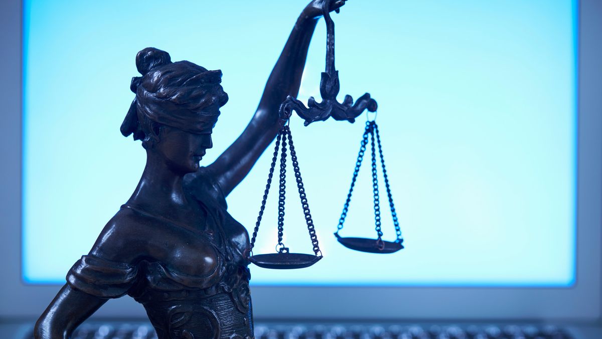 A statue of Lady Justice, a blindfolded woman raising two scales in the air to represent the legal system, set against a blurred background of a laptop with blue light streaming from the monitor, to represent the Computer Misuse Act.