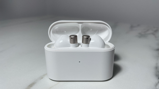 White and silver 1More PistonBuds Pro Q30 earbuds sitting in the charging case with the lid open. The case is sitting on a white marble-effect desk.