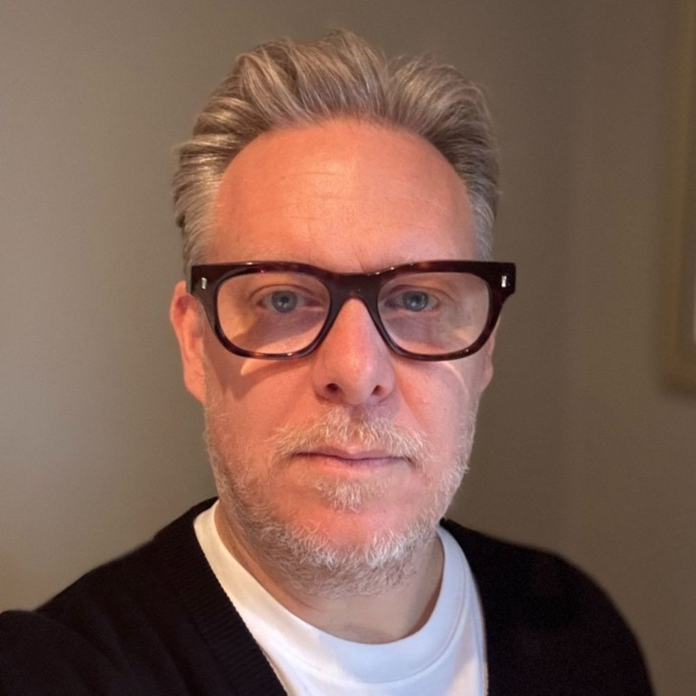 A man with short, neatly styled grey hair and dark-framed glasses looks directly at the camera. He wears a white T-shirt and dark cardigan.