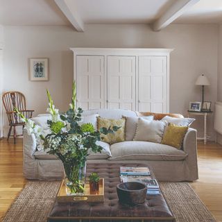 A large living room with a cream-coloured linen sofa with cushions, a leather ottoman-style coffee table with a vase of flowers and a vintage decorative wooden bowl