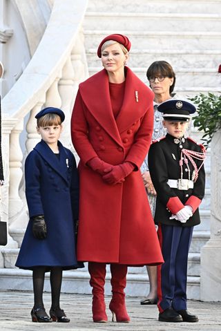 Princess Charlene of Monaco wears a bright red coat with matching stiletto boots, red gloves, a red dress, and a red hat, while she has blonde cropped hair, as she stands with her twins