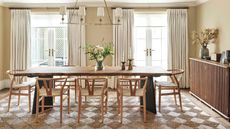 modern dining room with long rectangular timber dining table with wishbone chairs, checkered jute rug, two french doors with white curtains, cream walls, and a long fluted timber console along one wall styled with vase, speaker, and pictures