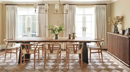 modern dining room with long rectangular timber dining table with wishbone chairs, checkered jute rug, two french doors with white curtains, cream walls, and a long fluted timber console along one wall styled with vase, speaker, and pictures