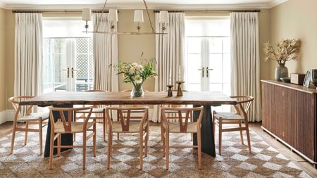 modern dining room with long rectangular timber dining table with wishbone chairs, checkered jute rug, two french doors with white curtains, cream walls, and a long fluted timber console along one wall styled with vase, speaker, and pictures