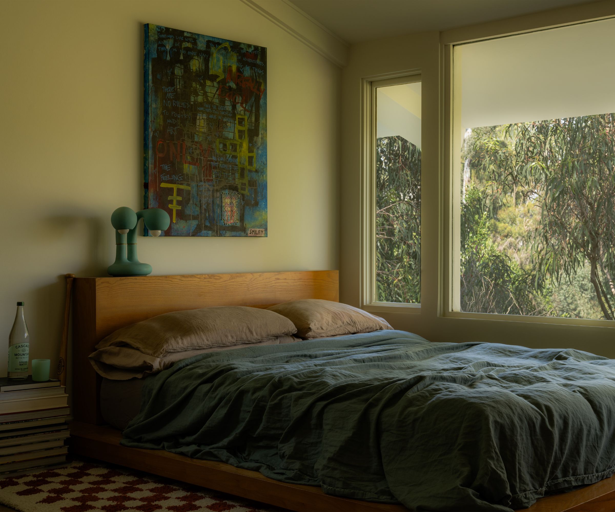 Wooden bed pushed up against a large window with blue bedding and brown pillowcases. Green walls, abstract artwork above bed, pile of books next to bed