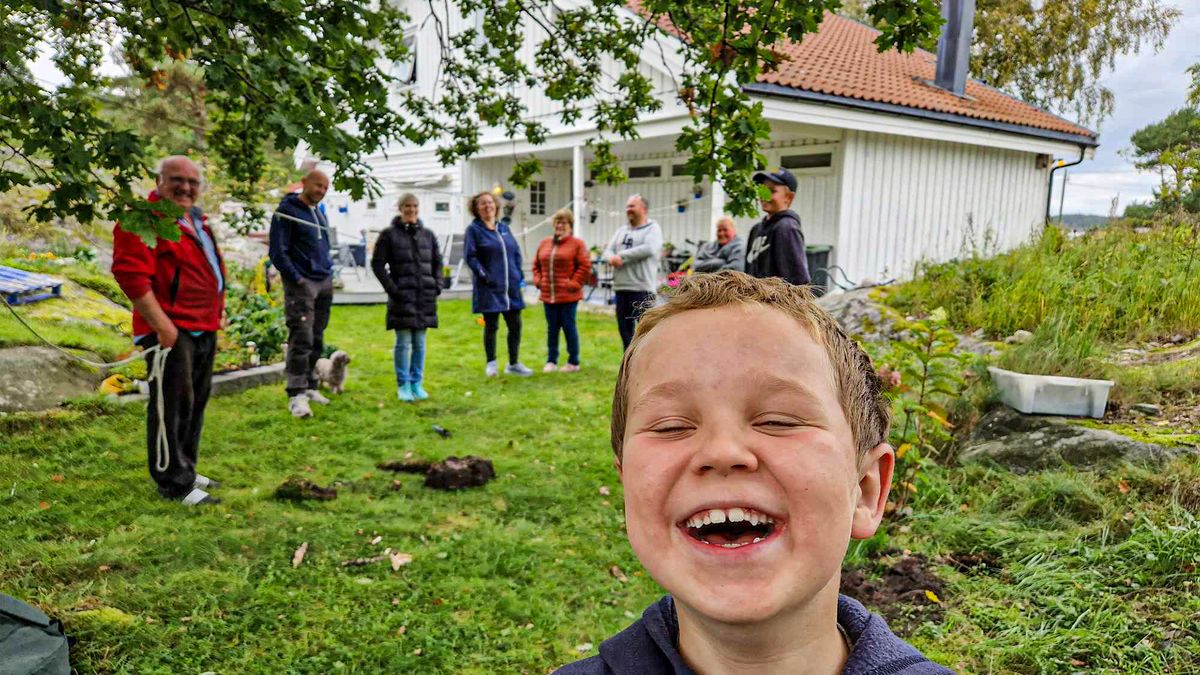 A family of all ages, including a young boy, smile in the green yard before a white house with a red roof.