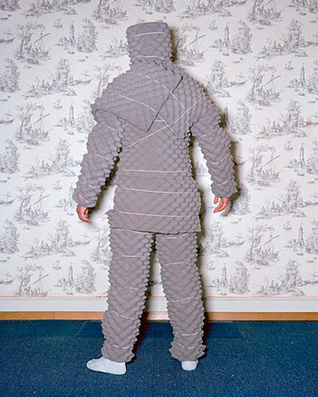A man wrapped in a grey fabric studded suit standing on a blue carpet in front of a wall with a plant patterned wallpaper on it.