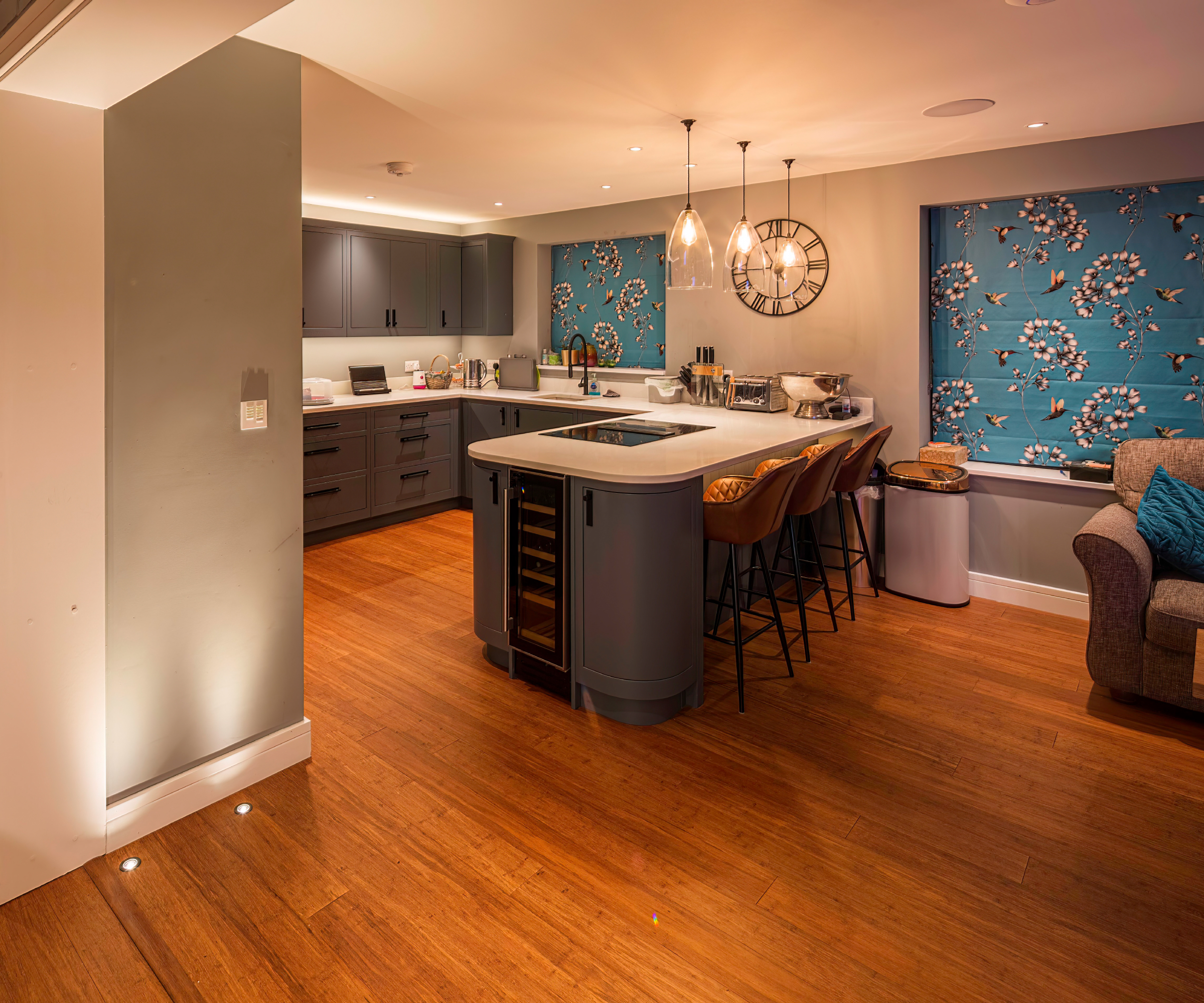 kitchen with grey units, wooden floor, blue and white floral blinds, glass pendant lights, spotlights and white multi circuit switch on wall