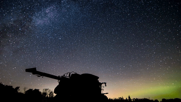 a silhouette of a large farming vehicle against a backdrop of stars and the milky way with northern lights glowing green in the distance.