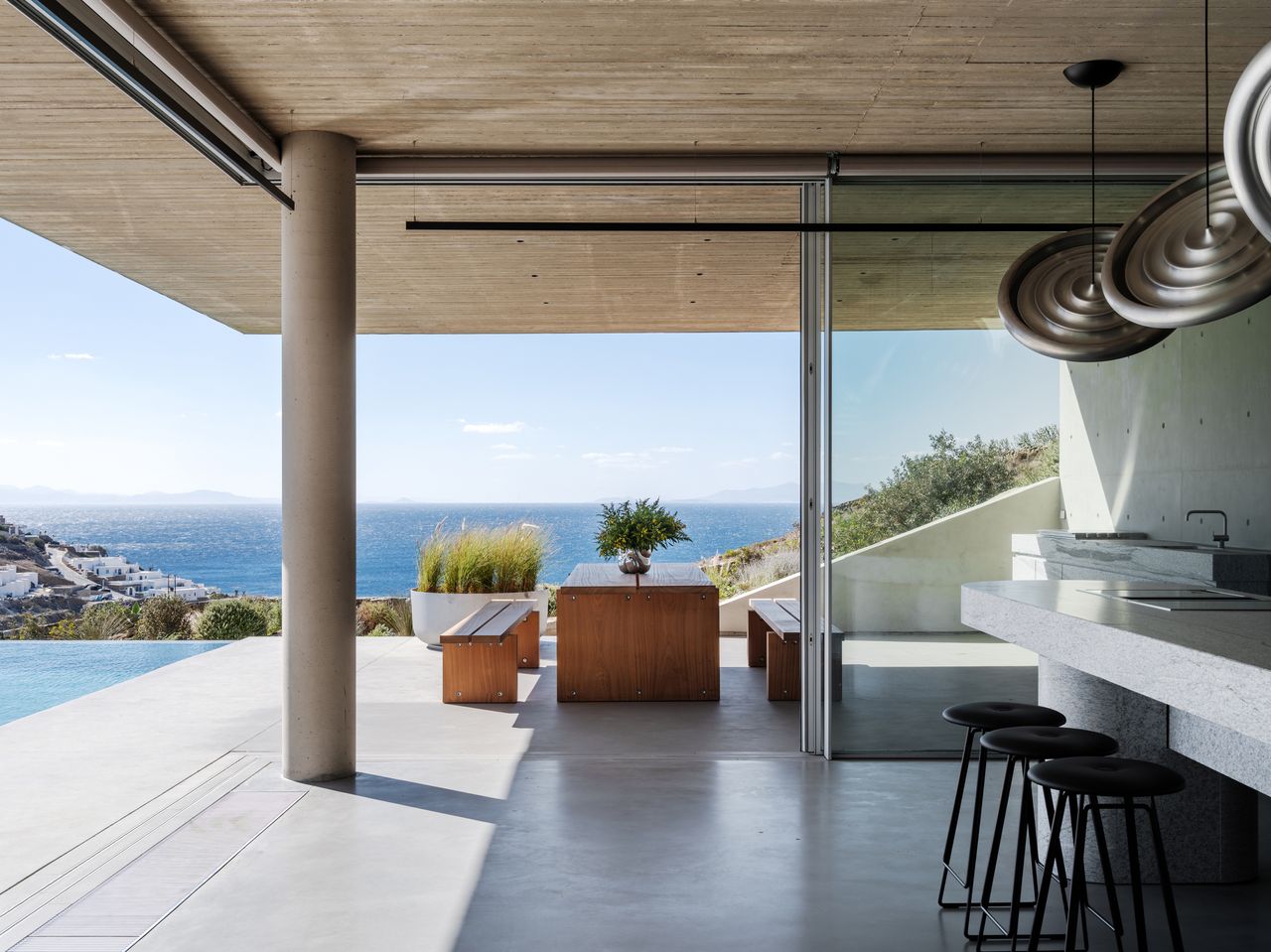brutalist kitchen in modern home with indoor-outdoor connection, timber dining table outside, swimming pool, concrete kitchen island, and three bar stools