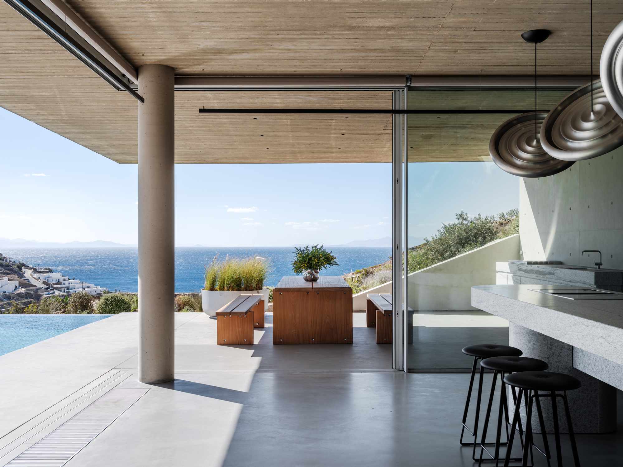 brutalist kitchen in modern home with indoor-outdoor connection, timber dining table outside, swimming pool, concrete kitchen island, and three bar stools