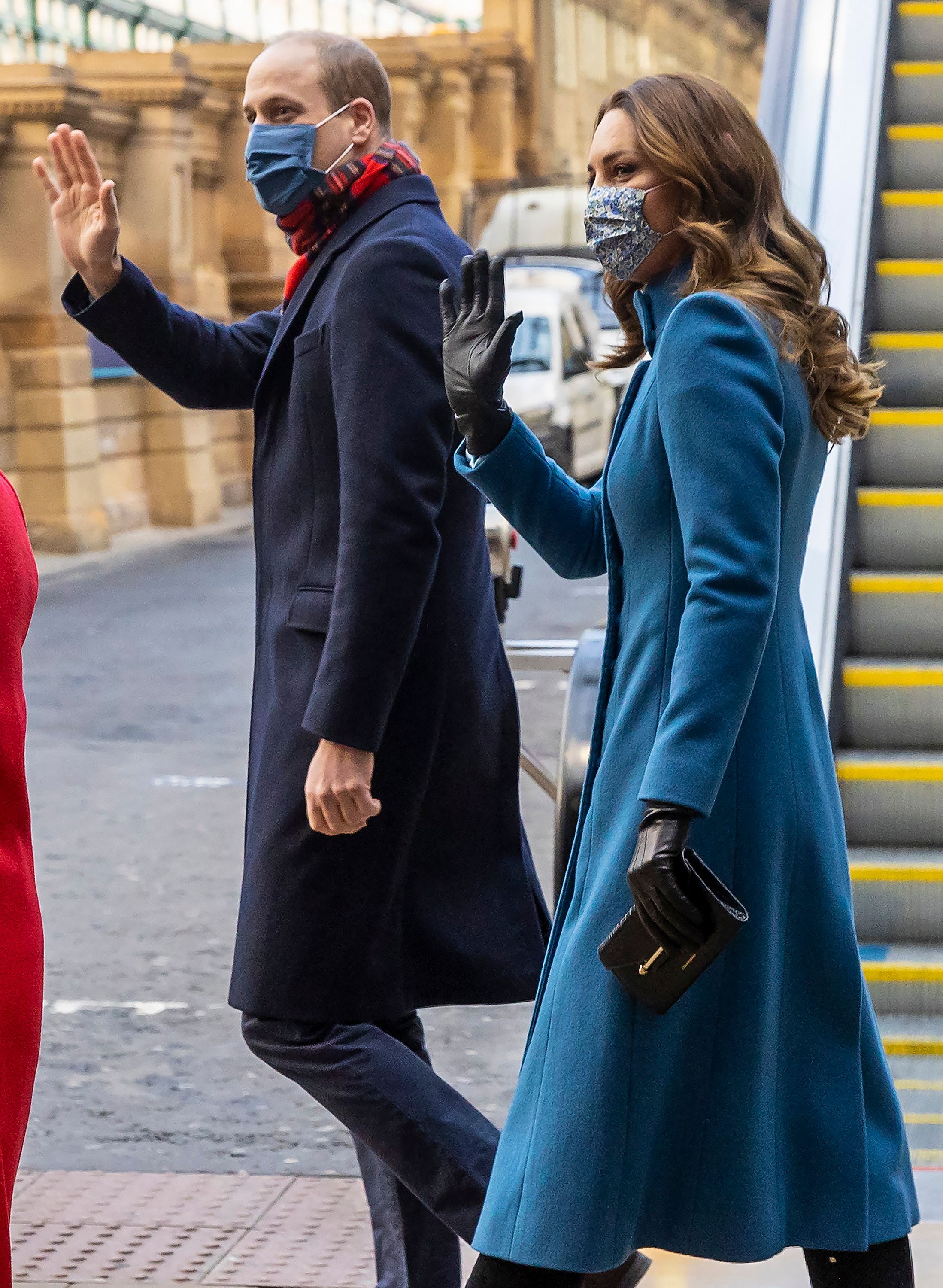 Princess Kate carrying a black clutch and wearing a blue coat waving next to Prince William, both in face masks