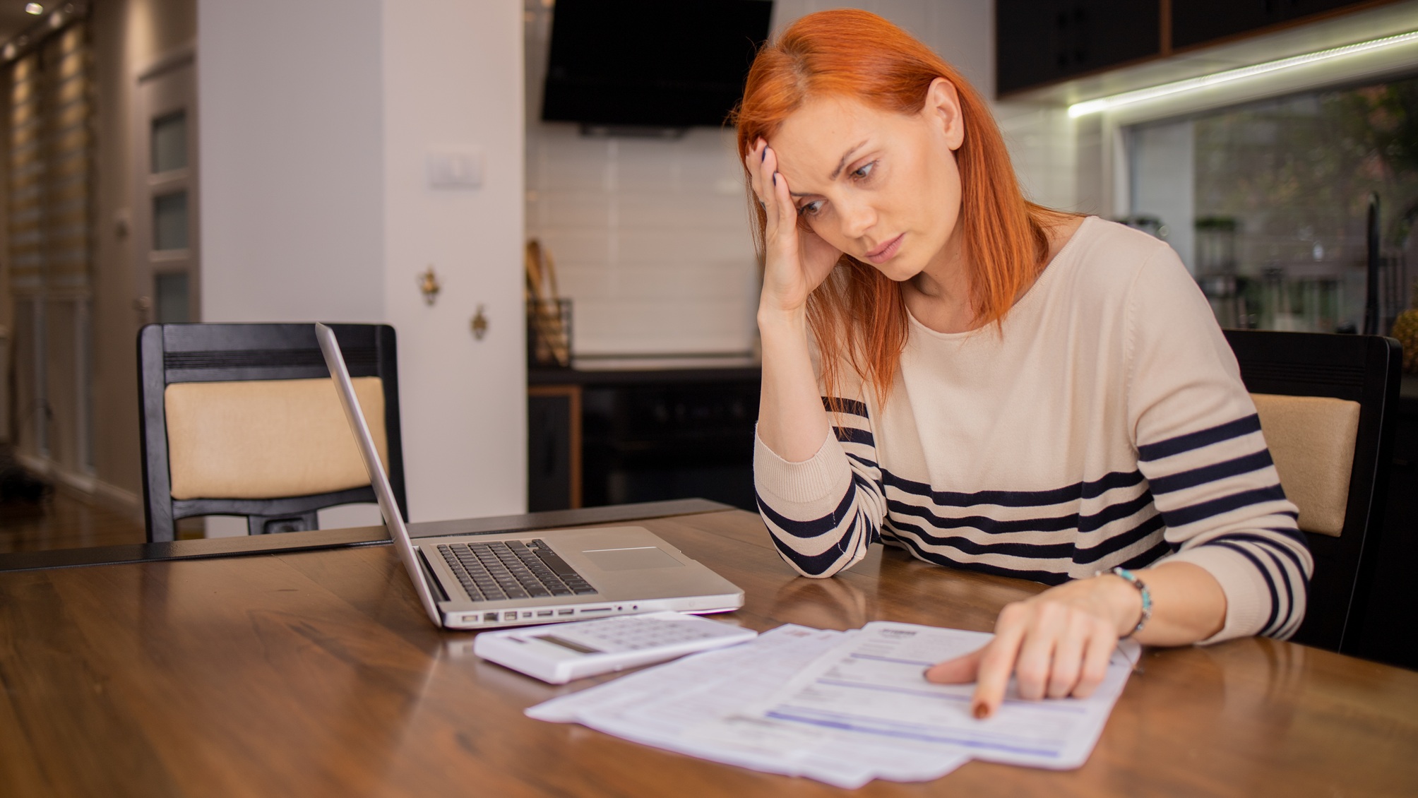 An older woman looks stressed as she looks over financial paperwork in her kitchen.