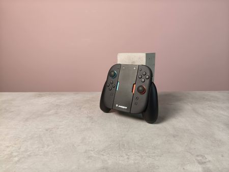 Snakebyte Ergo Grip S2 with Joy-Con 2 controllers attached, leaning on a plinth on a desk with pink wall in background